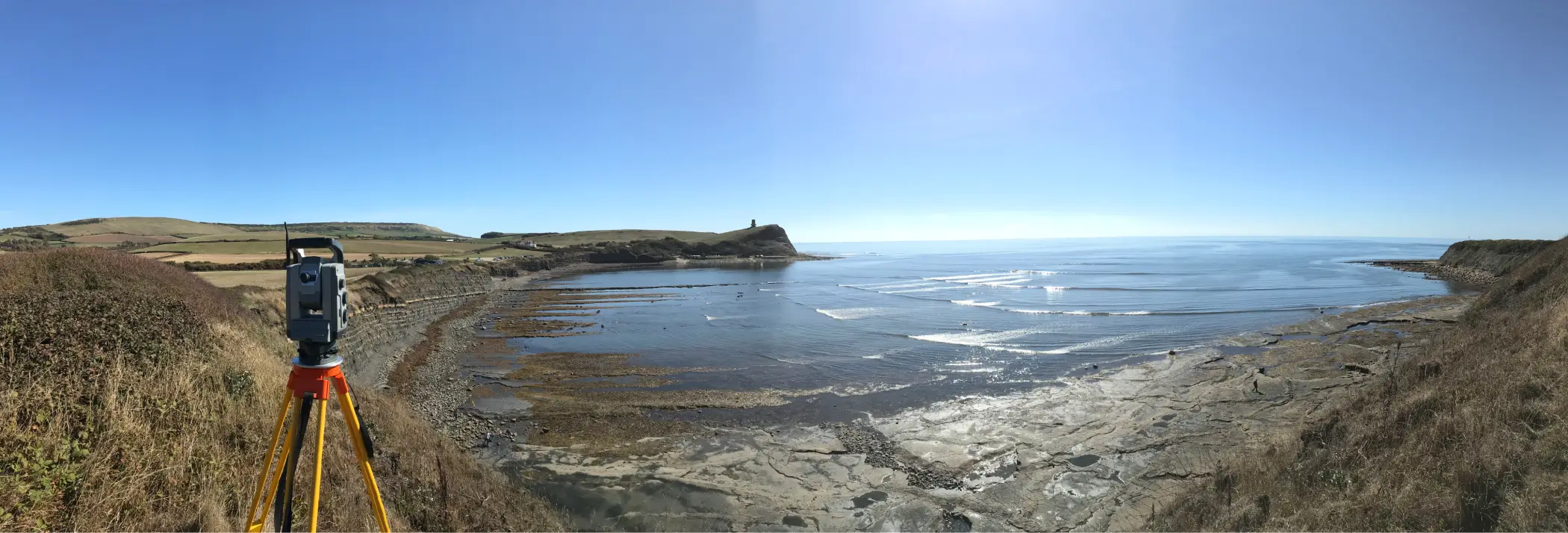 Land surveyors conducting coastal terrain analysis using a total station on a sunny day, overlooking the sea.