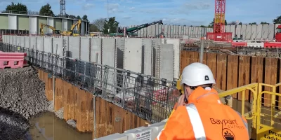 Land surveyor conducting measurements on a construction site with heavy machinery and retaining walls in the background.