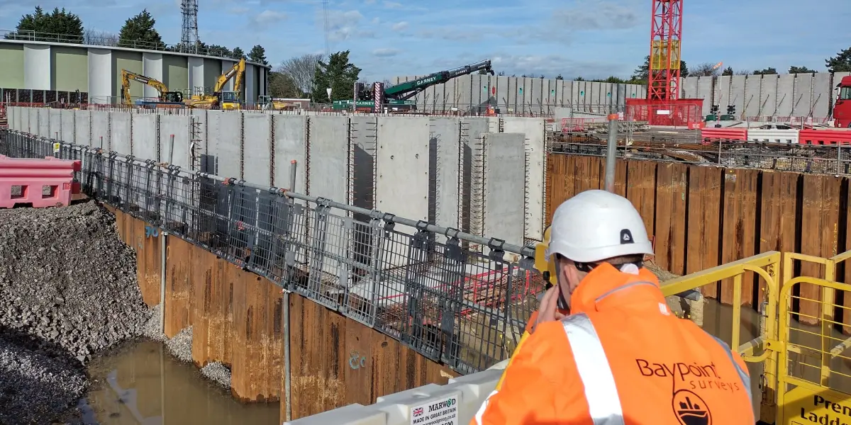 Land surveyor conducting measurements on a construction site with heavy machinery and retaining walls in the background.