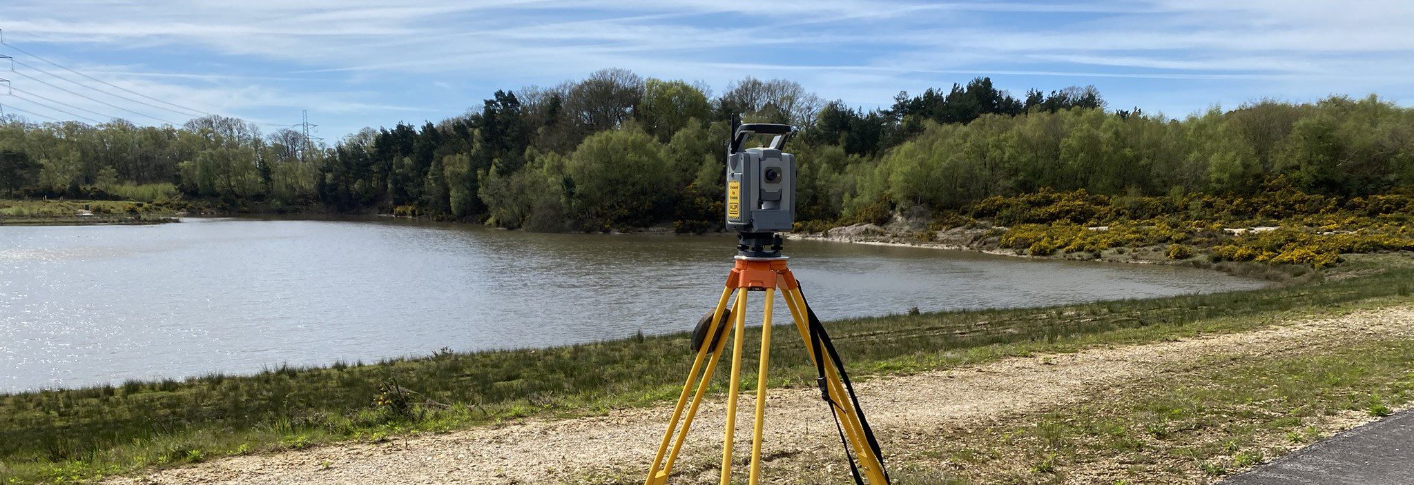 Land surveyor using equipment by a lake, conducting measurements in a scenic area with trees and clear skies.