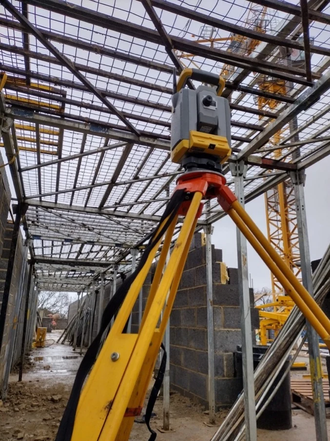 Laser scanning device on a tripod inside a construction site with steel framework and cranes in view.
